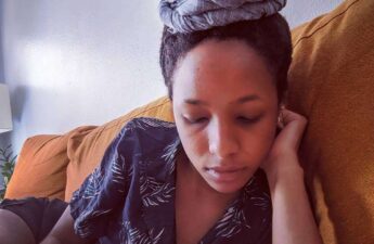 Black woman on orange couch looking down a notebook with hair up on a high bun with denim blue hair wrap
