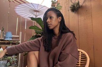 Photo for article - how to remove buildup in your locs once and for all. woman in brown sweater sitting on rattan chair on patio with fiddle fig plan in the background. Pink umbrella also in background. Woman has locs and is looking out of frame.
