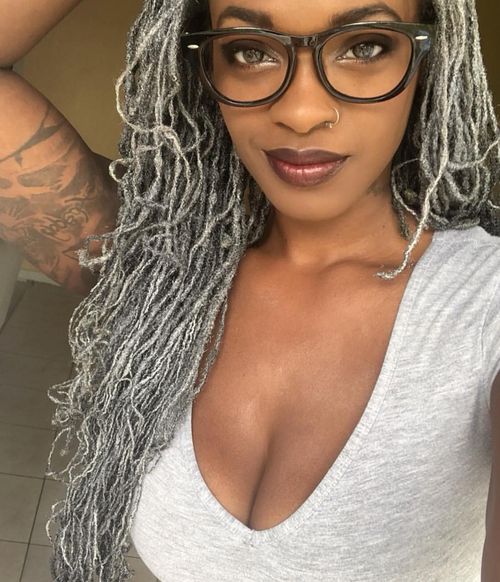 Woman with silver grey locs looking at camera wearing grey shirt. she has tattoos on her left arm, which is resting on her head.